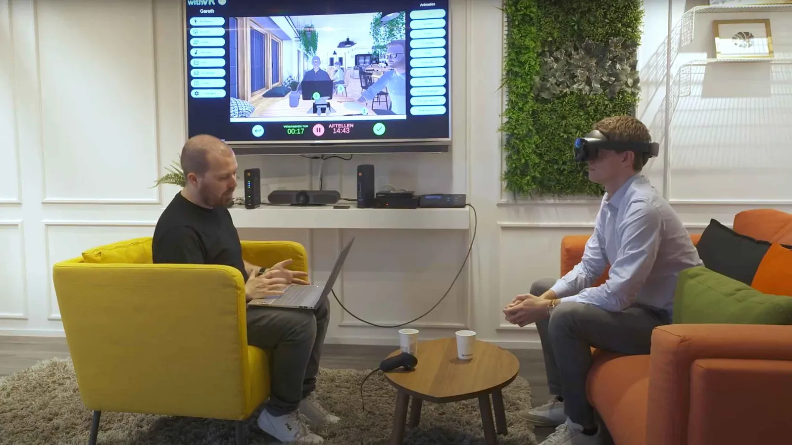 A young person wearing a withVR-branded headset while a therapist controls the virtual speaking situation from a laptop in the background